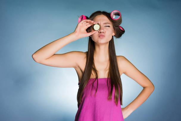 a funny young girl prepares for a party, on the head curlers for curling hair, holds a piece of cucumber to moisturize the skin of the face - Foto, Imagem
