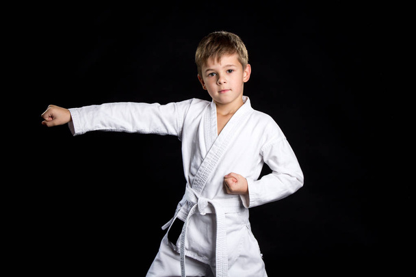 Straight karate arm hit. Kid in brand new kimono looking on the camera on isolated black background - Photo, Image