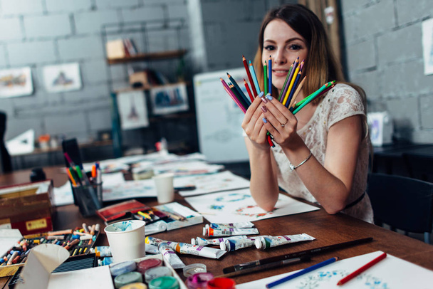 Mosolygó fiatal nő, egy csomó-ból ülő-on iskolapad, körülvéve művészeti ceruzák gazdaság kellékek - Fotó, kép
