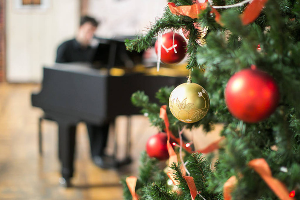 music, holidays, feast concept. close up of marvelous sparkling balls of golden and crimson colours, that hang on the christmas tree with red ribbons on the branches on the background with pianist - Photo, Image