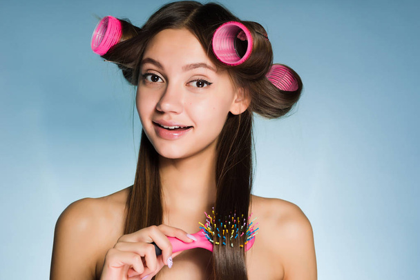 lovely young girl makes a beautiful fashionable hairstyle with the help of large curlers, combs her hair - Foto, imagen