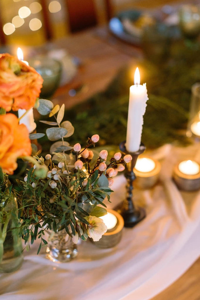 dating, hospitality, comfort concept. tall slim candle that is standing in black holder surrounded by lots of small ones, placed by it on the table with romantic setting of flowers and dishes - Fotoğraf, Görsel