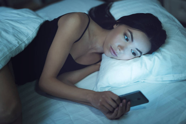 La chica está en un cuarto oscuro en la cama. Mira el smartphone. Influencia de las redes sociales en la juventud
. - Foto, imagen