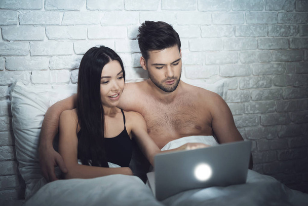 Girl is lying in a dark room in bed with a guy. A guy with a girl in bed is looking at the laptop. - Φωτογραφία, εικόνα