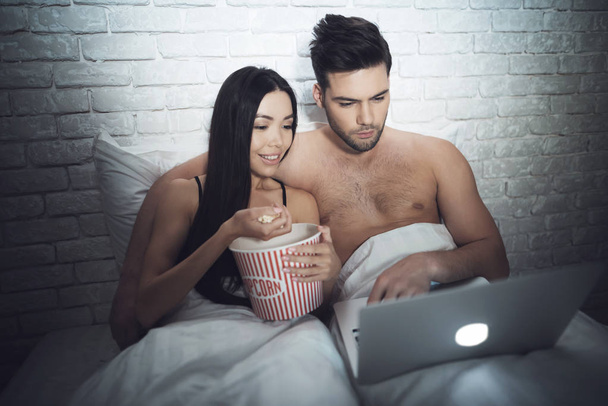 Girl is lying in a dark room in bed with a guy. A guy with a girl in bed is looking at the laptop. - Фото, изображение