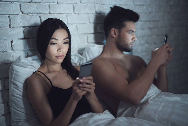 Chica con chico yace en un cuarto oscuro en la cama. Están mirando el teléfono inteligente. Influencia de las redes sociales
. - Foto, imagen