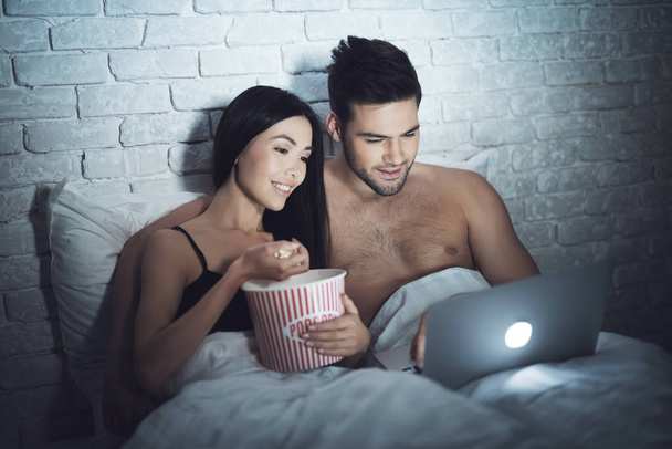 Girl is lying in a dark room in bed with a guy. A guy with a girl in bed is looking at the laptop. - Foto, Bild