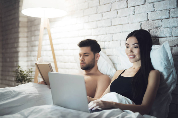 Girl and guy are in bed. The guy reads the book before going to sleap. Girl working on laptop in bed. - Fotografie, Obrázek