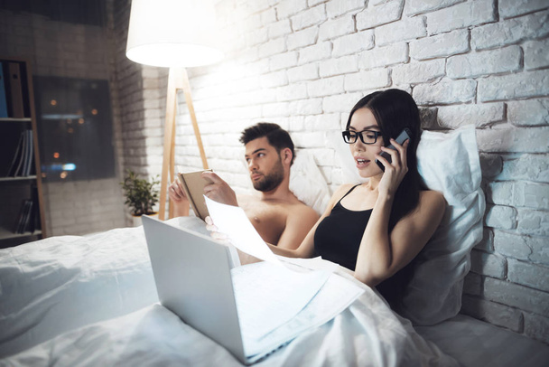 Girl and guy are in bed. The guy reads the book before going to sleap. Girl working on laptop in bed. - Fotoğraf, Görsel