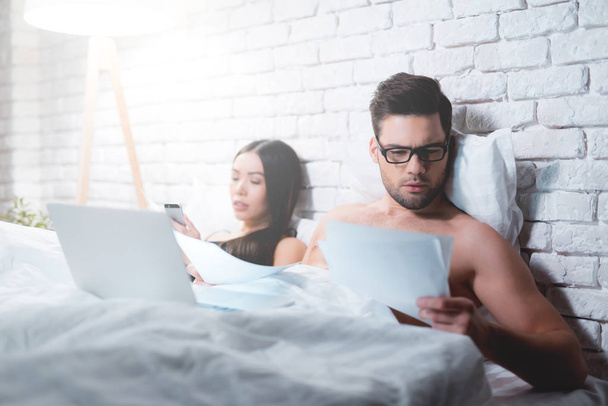 A guy is lying in bed and working on a laptop. The guy does not pay attention to his girlfriend. - Foto, Bild