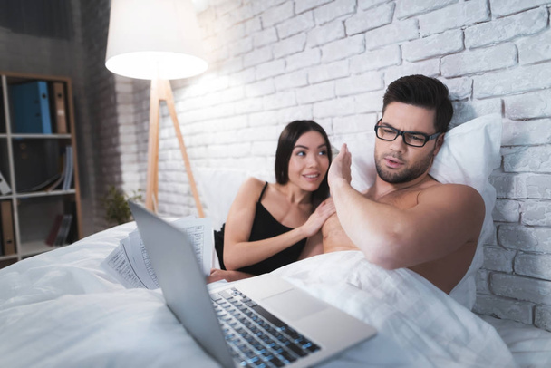 A guy is lying in bed and working on a laptop. The guy does not pay attention to his girlfriend. - Photo, Image
