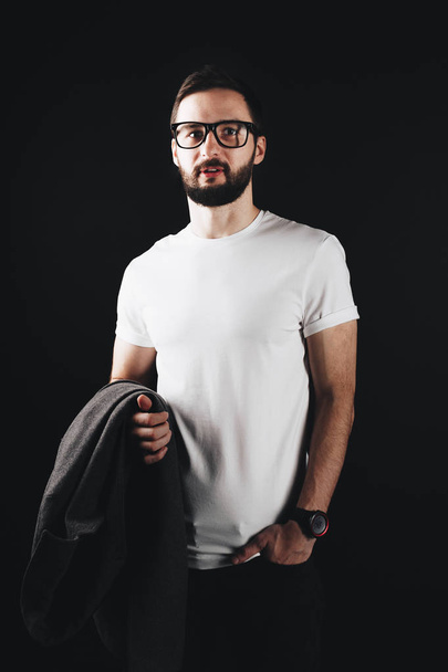 Handsome bearded man wearing glasses and white t-shirt and holds in his hand a coat on a black background - Φωτογραφία, εικόνα