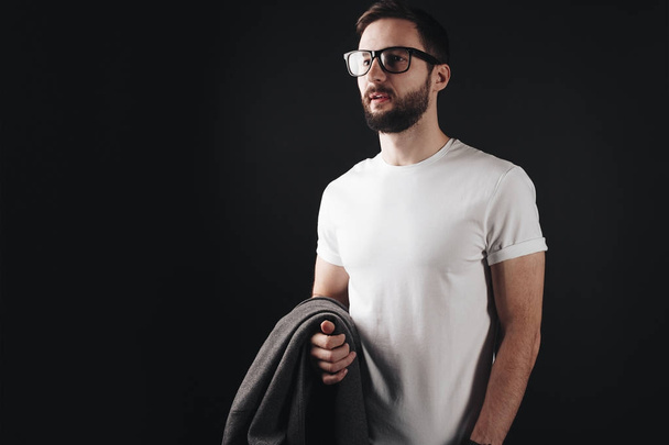 Young bearded pensive business man in glasses wearing blank white t-shirt and holds coat on black background - Fotografie, Obrázek