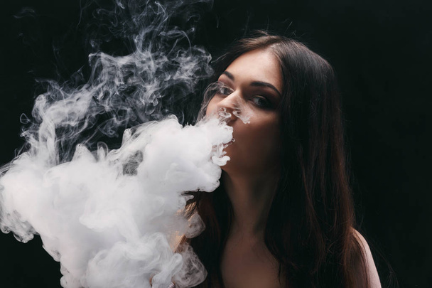 Young woman vaping e-cigarette with smoke on black - Foto, imagen