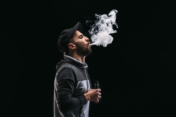 Young man vaping e-cigarette with smoke on black - Фото, изображение