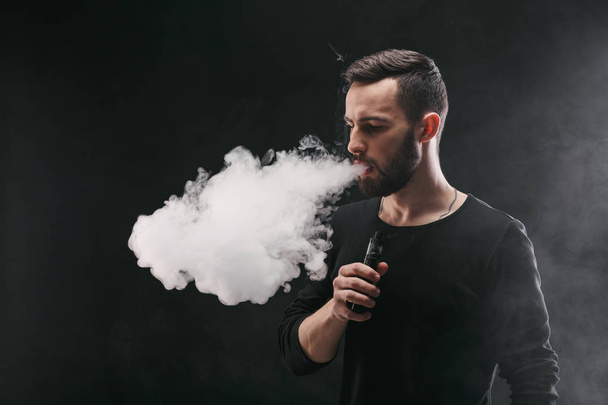 Young man vaping e-cigarette with smoke on black - Foto, immagini