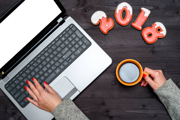 Top view of woman hand holding cup of hot coffee and typing on open laptop keyboard with blank white screen, copy space. New year 2018 sign symbol from gingerbread cookies, free space. Flat lay - Foto, Bild