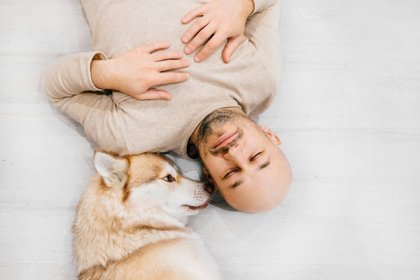 Adult bald man with husky puppy sleeping on floor. Owner with pet together at home. Kind and soulful emotions. Lovely dog resting with young male. Guy with beloved domestic animal hugging each other. - Фото, изображение