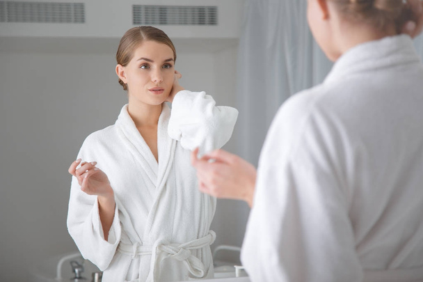 Confident young woman in bathrobe looking at mirror - 写真・画像