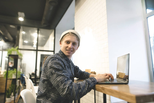 A positive student works at a computer in a cozy cafe and looks at the camera. Hipster frees up at the computer. Work at a computer in a cafe. - Photo, Image
