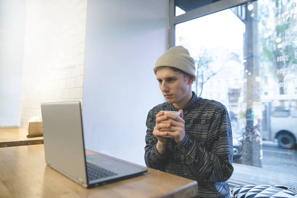 Молодий чоловік сидить у затишному кафе з чашкою кави в руках і дивиться відео на ноутбук. Стильний студент за комп'ютером у кафе
. - Фото, зображення