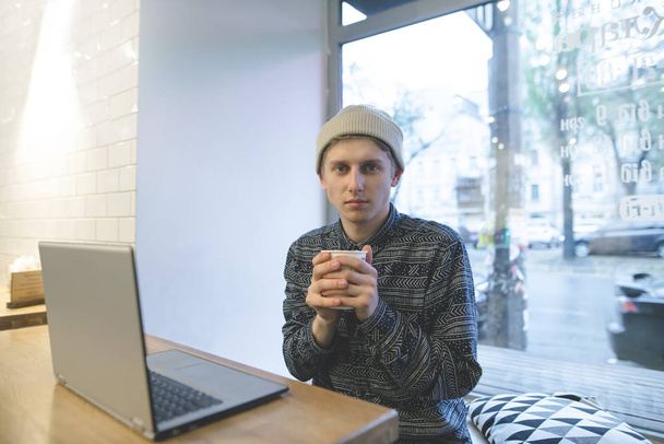 Un joven hombre de hip-hop se sienta en un acogedor café cerca de una computadora portátil y calienta el café. El freelancer trabaja en una cafetería en la computadora y bebe café
. - Foto, imagen