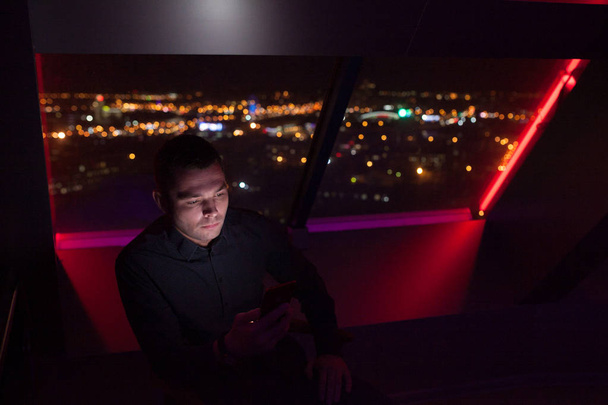 Businessman uses a smartphone sitting by the window in the office on a background of a night city. - Zdjęcie, obraz