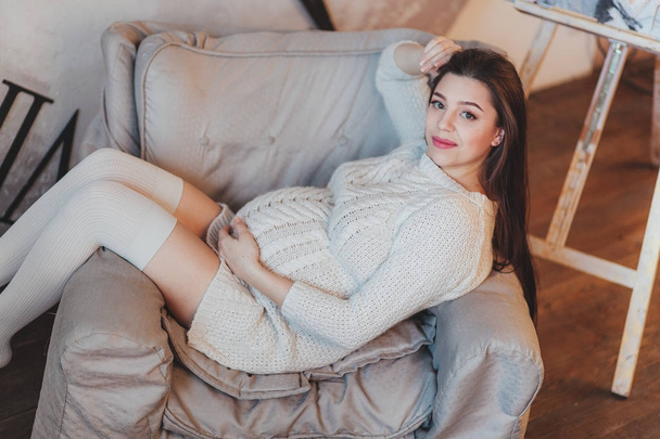 Pregnant brunette girl in a white knitted sweater lying in armchair - Foto, imagen
