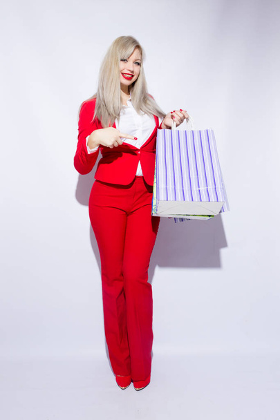 Image of young blonde lady in red costume posing with shopping bags and looking to camera over white background - Photo, Image
