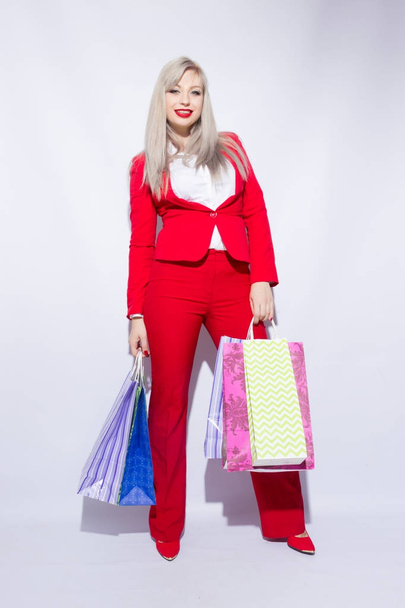 Image of young blonde lady in red costume posing with shopping bags and looking to camera over white background - Foto, immagini