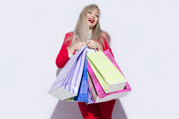 Image of young blonde lady in red costume posing with shopping bags and looking to camera over white background - Fotografie, Obrázek