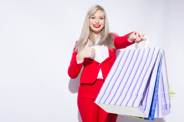 Image of young blonde lady in red costume posing with shopping bags and looking to camera over white background - Zdjęcie, obraz