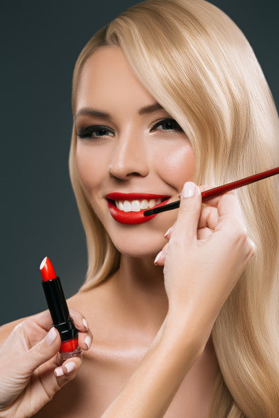 Portrait of beautiful blonde hair girl with visagiste hands applying lipstick with brush, isolated on black - Photo, Image