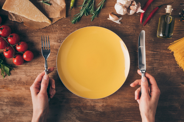 vista cortada de mãos com faca e garfo na placa com massa linha e ingredientes frescos em torno de mesa de madeira
 - Foto, Imagem