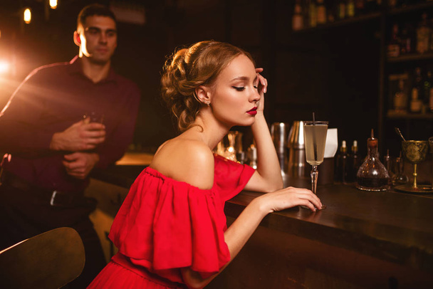 attractive young woman in red dress sitting at bar counter, handsome man on background. Date in nightclub, attractive couple in pub  - Fotoğraf, Görsel