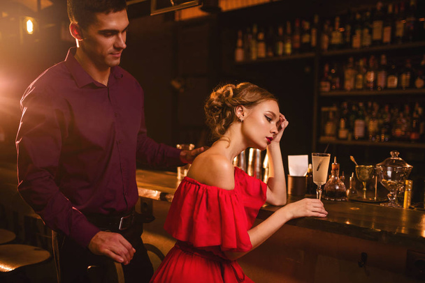 aantrekkelijke jonge vrouw in rode jurk zit aan toog, knappe man achter haar. Datum in nachtclub, aantrekkelijk koppel in pub  - Foto, afbeelding