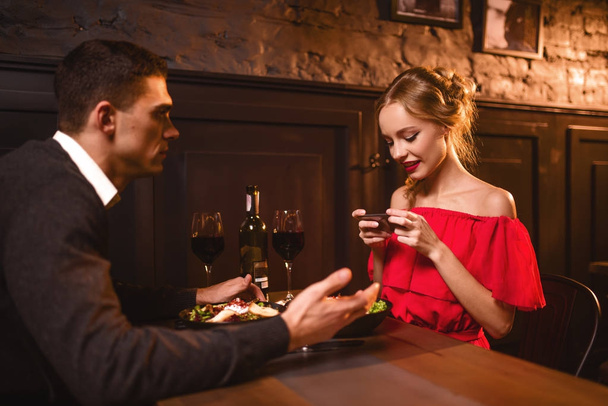 Uśmiechnięta kobieta w czerwonej sukience biorąc zdjęcie jedzenie na telefon aparat. Młoda miłość para w restauracji, romantyczną randkę. Elegancka Kobieta w czerwonej sukience i przystojny mężczyzna jadalnia  - Zdjęcie, obraz