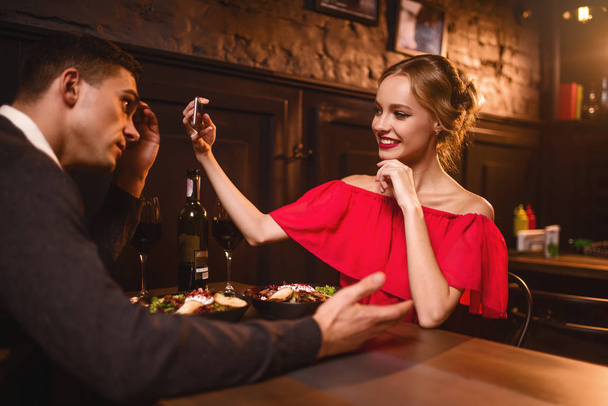 Gülümseyen kadın erkeğini fotoğraf telefon kamera çekici Kırmızı elbiseli. Gençlik aşkları iki restoran sonra romantik tarih. Kırmızı elbise ve yakışıklı adam yemek zarif kadın  - Fotoğraf, Görsel