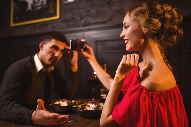 携帯電話のカメラで彼女の男の写真を取っている赤いドレスを着た女性の笑みを浮かべてください。若い愛カップルのレストランでロマンチックな日付。赤いドレスとハンサムな男がダイニングで優雅な女性  - 写真・画像