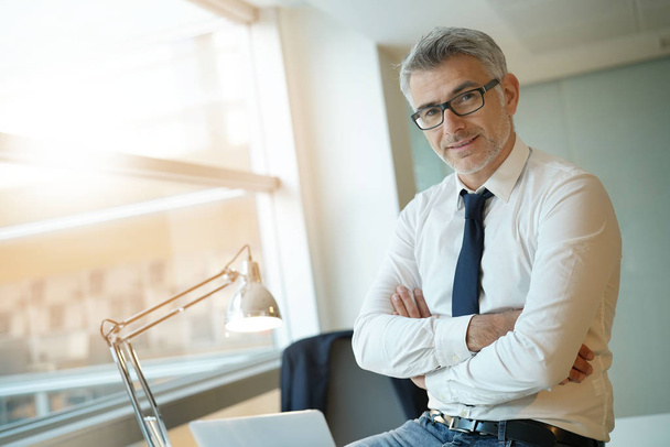 Confident businessman sitting on desk with arms crossed - Фото, изображение