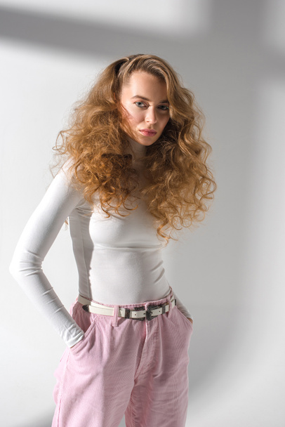 confident stylish girl with curly hair standing with hands in pockets and looking at camera - Foto, immagini