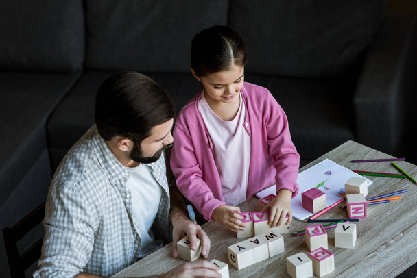 father with daughter sitting at table and making words by cubes with letters at home  - Photo, Image