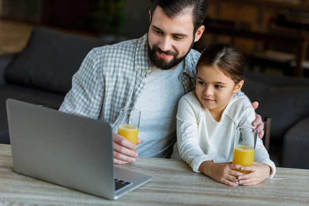 smiling father with daughter sitting at table with juice and using laptop - Фото, изображение