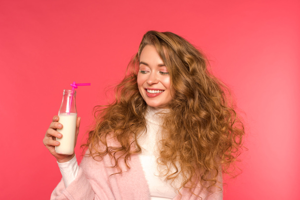 smiling girl looking at bottle with milkshake and plastic straw isolated on red - Foto, imagen
