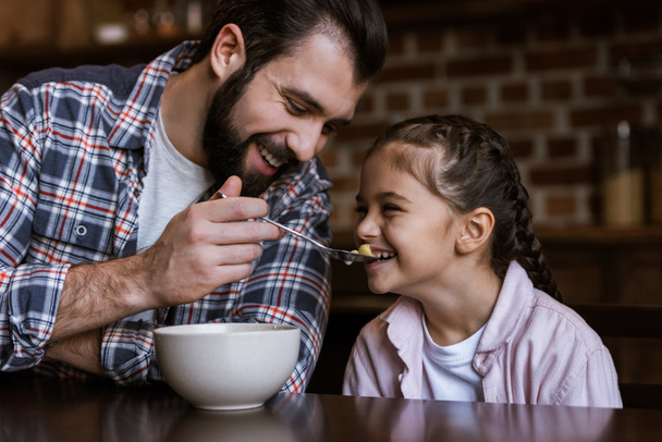 família alegre à mesa, pai alimentando filha por lanches com leite na cozinha
 - Foto, Imagem