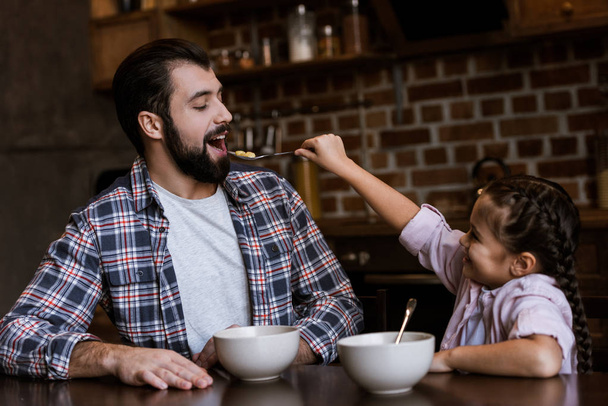 cheerful family at table, daughter feeding father by snacks with milk at kitchen - Photo, Image