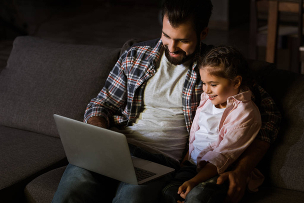 cheerful father with daughter sitting on couch and using laptop - Photo, image