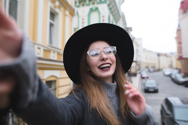 Elegante ragazza con l'apparecchio prende selfie krasvoho sullo sfondo del paesaggio urbano. La ragazza turista sullo sfondo fa selfie belle strade del centro storico
. - Foto, immagini