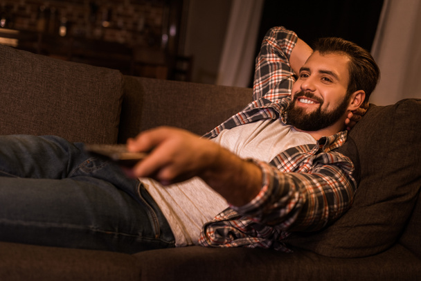 νεαρός άνδρας θέτοντας στον καναπέ και βλέποντας τηλεόραση στο σπίτι - Φωτογραφία, εικόνα