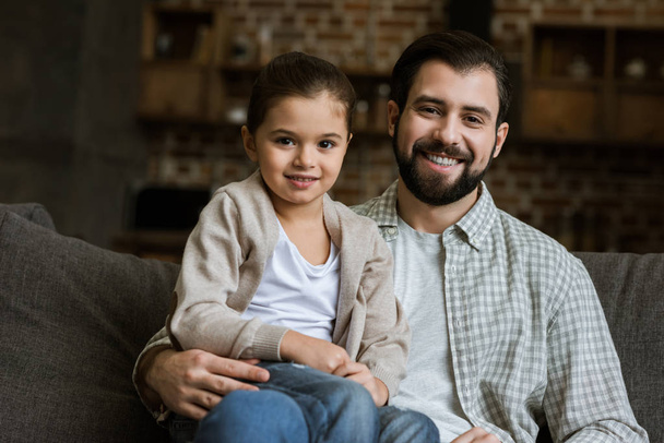 happy father and daughter hugging on couch and looking at camera at home - Фото, изображение